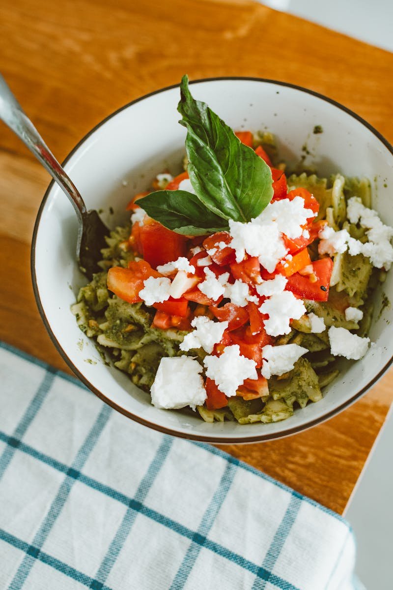 Delicious pesto pasta topped with fresh tomatoes, feta cheese, and basil in a white bowl.