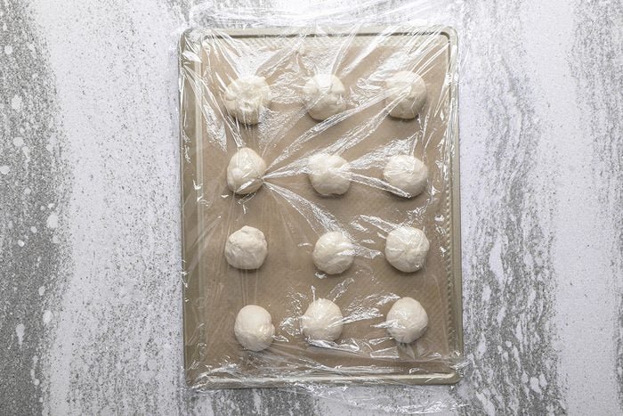 A baking sheet with twelve dough balls covered in plastic wrap, set on a marbled countertop.